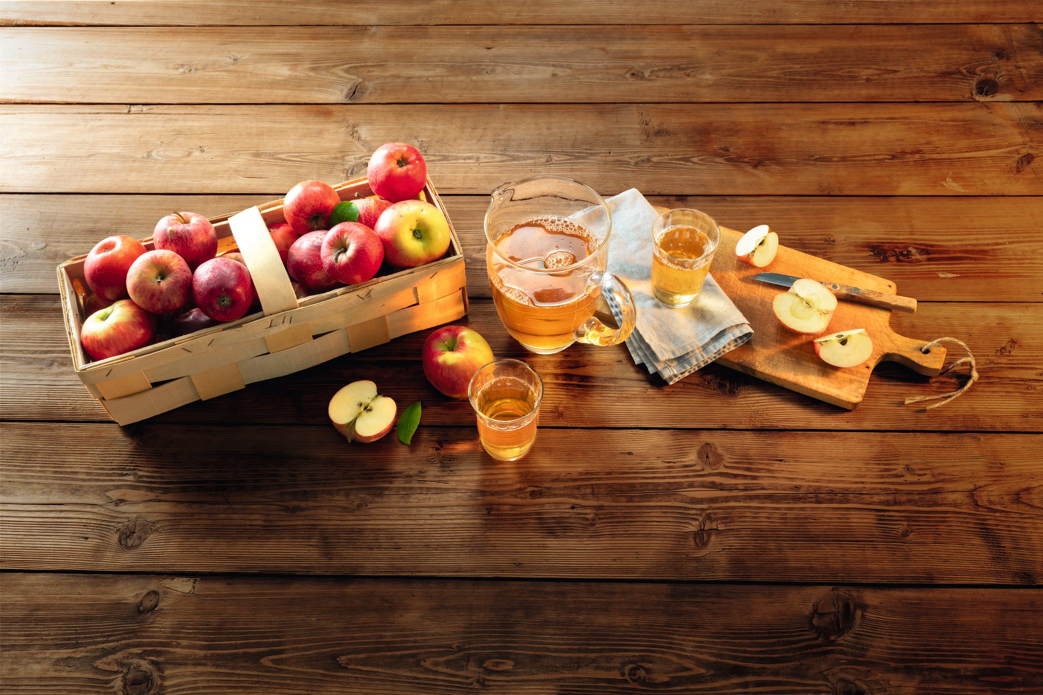 Ein Holzkorb mit Äpfeln. Daneben steht eine Karaffe mit Apfelsaft und ein Glas Apfelsaft. Ein Holzbrett mit einem Messer und Apfelhälften. Ein ganzer und ein halber Apfel stehen neben dem Glas.